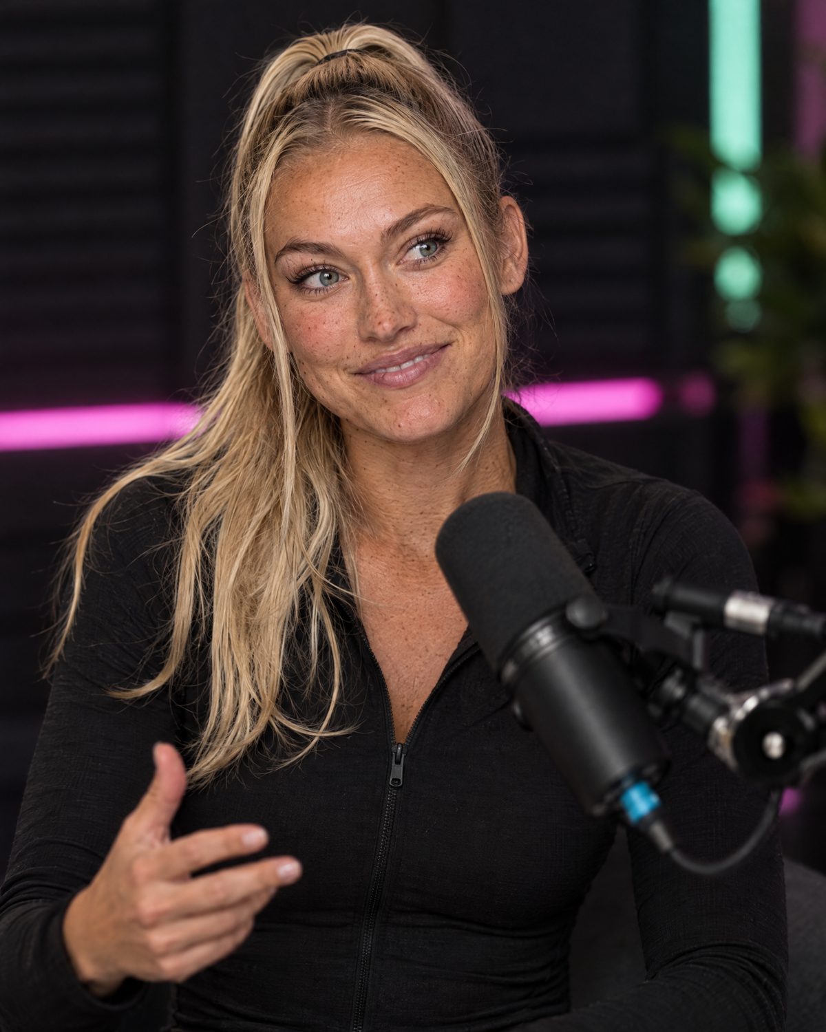 Nina Vellin mid-sentence in a dark podcast studio with pink and mint lighting.